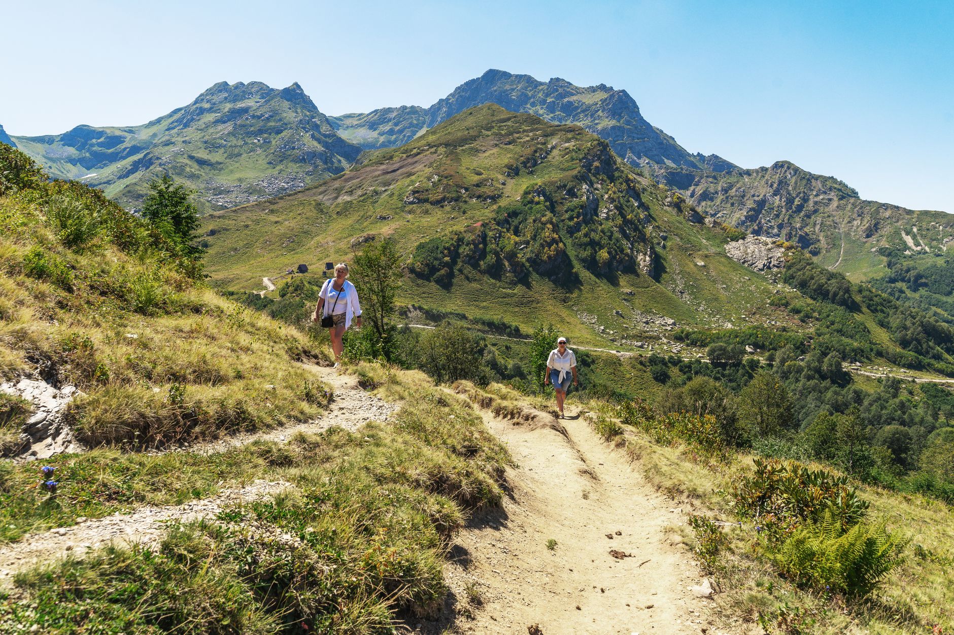 Deux randonneuses marchent sur un sentier de terre en montagne, avec des pentes herbeuses et des sommets rocheux sous un ciel bleu.